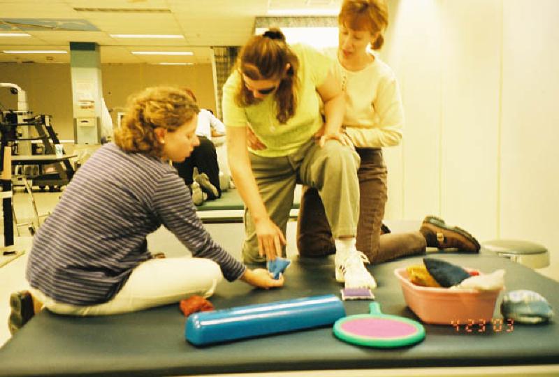 Peggy and Jean doing exercises with Marla's injured right hand and arm. Here, Marla is using the injured arm to pick up, move, and place bean bags on the therapy mat.